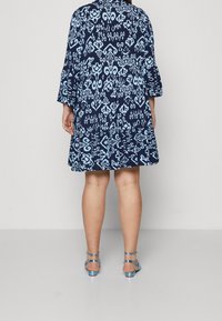 En person som står, iklädd en knälång mörkblå klänning med ljusblå abstrakta mönster och ljusblå ankelbandsandaler, vänd bort.