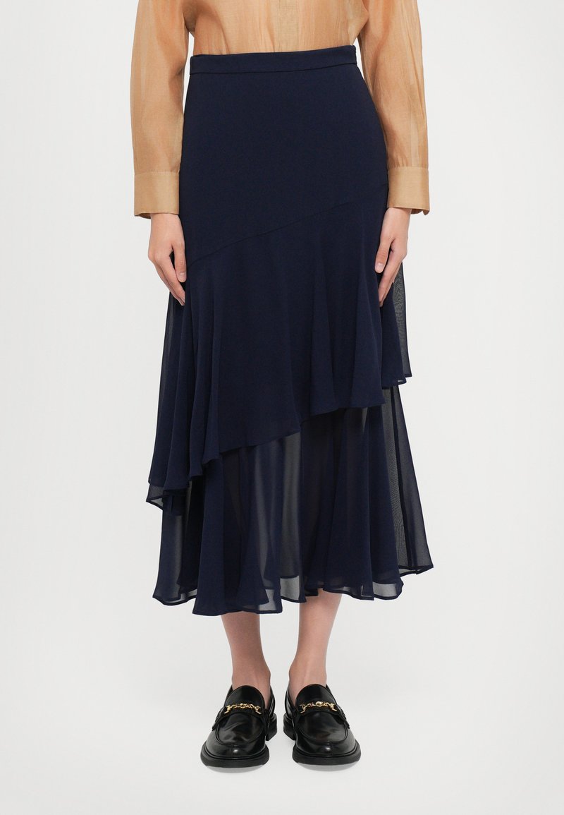 Femme portant une jupe midi en chiffon asymétrique bleu marine, un blouse à manches longues beige, et des mocassins en cuir noirs avec un détail en chaîne dorée.