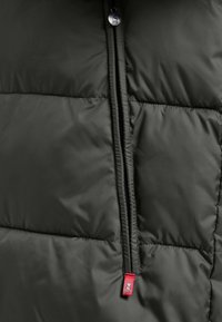 Donkergroene pufferjack met horizontale gestikte textuur, voorzien van een zwarte rits en een klein rood label met een embleem.