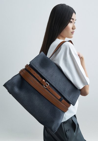 Ung kvinde med langt sort hår iført hvid skjorte og sorte bukser bærer en stor grå og brun læderbesat tote taske over skulderen.