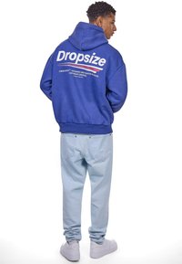 Niebieska bluza z kapturem z białym napisem "Dropsize" i logo na plecach, w połączeniu z jasnoniebieskimi dżinsami i białymi sneakersami.