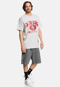 Graues Baumwoll-T-Shirt mit rotem grafischen Druck auf der Vorderseite, kombiniert mit grauen Denim-Shorts. Dazu gehören schwarze Socken und weiße Sneakers.