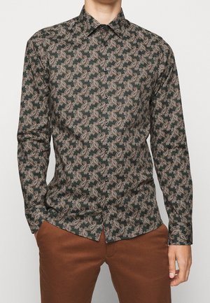 Mann trägt ein dunkelgrünes Langarmhemd mit braunem Paisleymuster und braune Hose, Hände in den Taschen, schaut nach vorne.
