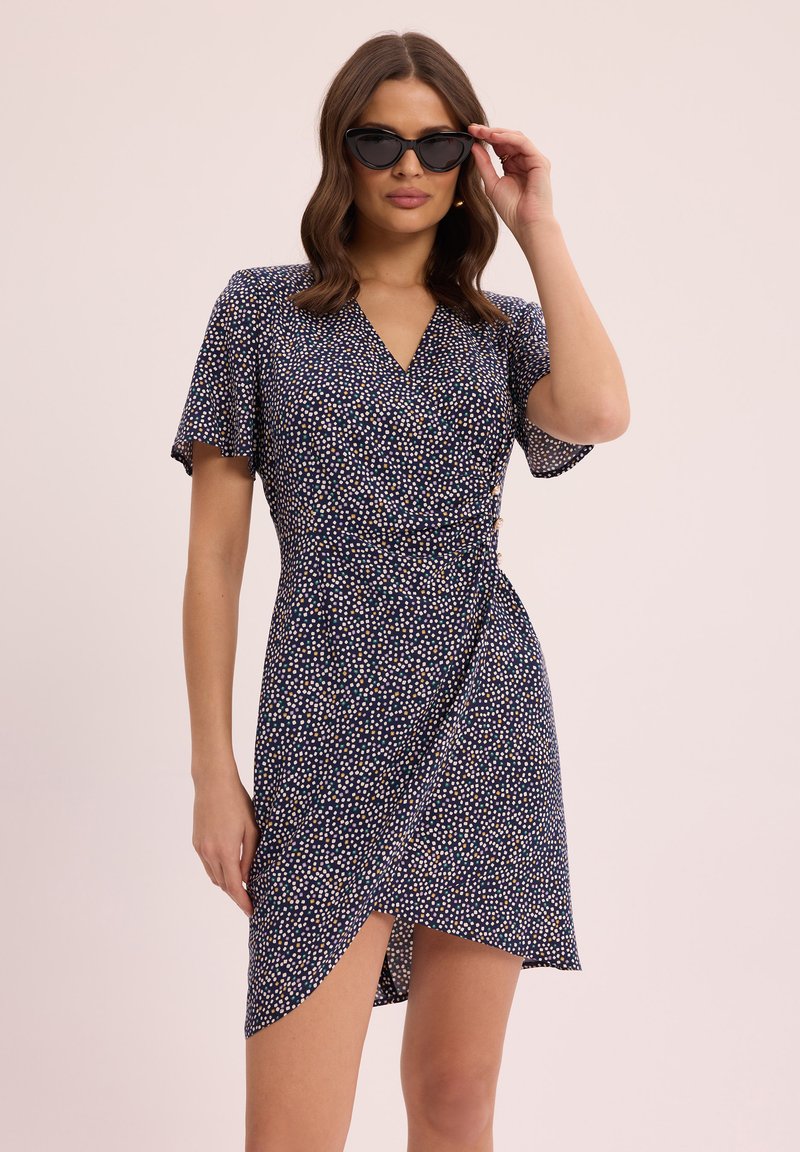 Niebieska kwiecista sukienka w stylu wrap z krótkimi rękawami, z dekoltem w kształcie litery V, we wzór w kropki oraz bocznym wycięciem. Czarne okulary typu cat-eye dopełniają stylizację.