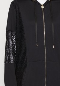 Černá mikina na zip s krajkovým panelem na levém rukávu, hladkým materiálem, dvěma předními kapsami a zlatými kovovými doplňky.