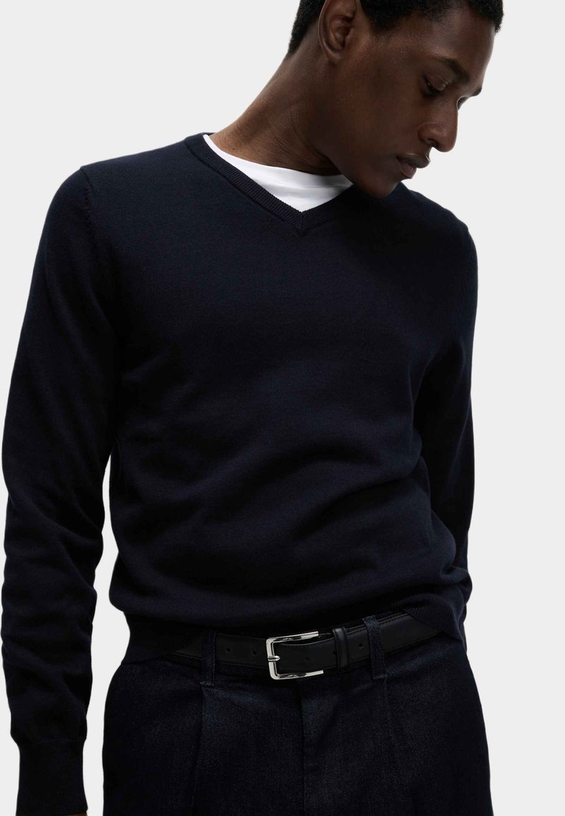 Mann trägt einen eng anliegenden schwarzen V-Ausschnitt-Pullover über einem weißen Unterhemd, schwarze Hose und einen schwarzen Gürtel mit silberner Schnalle, schaut nach unten.