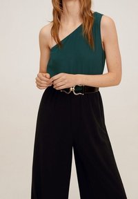Haut vert asymétrique à une épaule avec une texture lisse, associé à un pantalon noir large taille haute. Comprend une ceinture dorée avec une boucle décorative.
