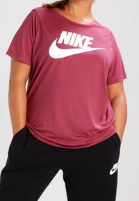 Hnědý Nike tričko s krátkým rukávem, zdobené velkým bílým logem Nike. Hladký materiál, volný střih, kombinováno s černými kalhotami.