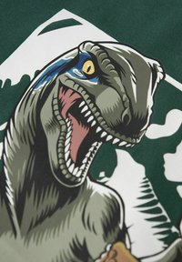 Grafische print van een dinosaurus op een groene stoffen shirt. De dinosaurus heeft grijstinten, een open mond, scherpe tanden en een blauwe accent.