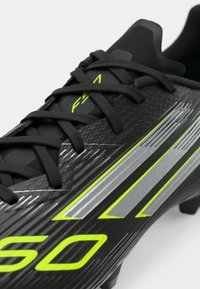 Schwarzer Fußballschuh mit strukturierter Oberfläche, ausgestattet mit grünen und silbernen diagonalen Streifen, schwarzen Schnürsenkeln und einer abgerundeten Zehenform.