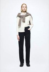 Duga majica s kremastim rukavima, crne uske hlače, smeđi pleteni džemper zavezan oko ramena, crne masivne čizme, jednobojna bijela pozadina.