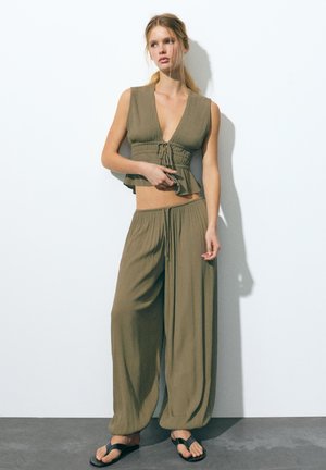 Jeune femme debout contre un mur, portant un crop top sans manches vert olive avec cordon de serrage et un pantalon ample assorti avec des sandales noires.