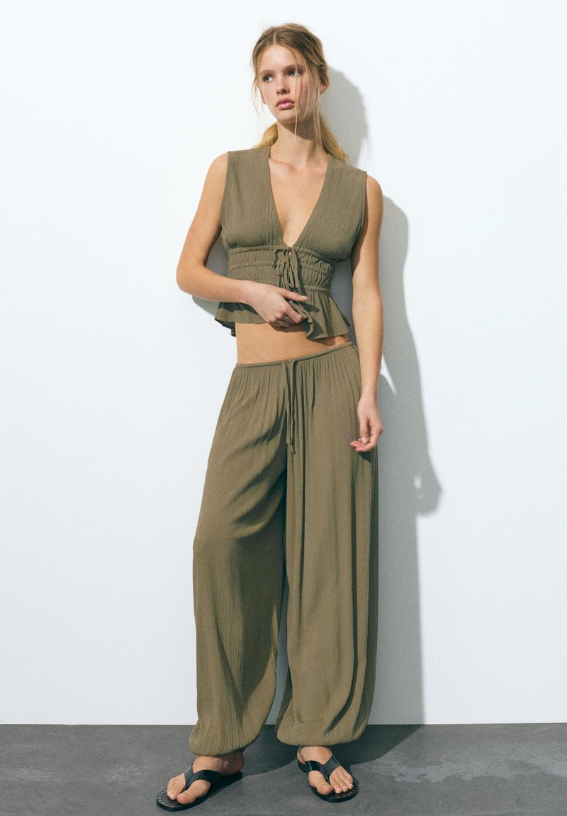 Jeune femme debout contre un mur, portant un crop top sans manches vert olive avec cordon de serrage et un pantalon ample assorti avec des sandales noires.