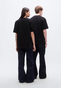 Μαύρα βαμβακερά T-shirts, άνετη εφαρμογή, συνδυασμένα με σκούρο μπλε, φαρδιά τζιν παντελόνια. Άτομα εμφανίζονται από πίσω, λιτός σχεδιασμός.