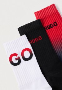 Τρία ζευγάρια κάλτσες: λευκές με κόκκινο και μαύρο κείμενο "GO", μαύρες με κείμενο "HUGO" και με διαβάθμιση από κόκκινο σε μαύρο με κείμενο "HUGO", με ριμπ υφή.