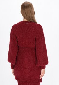 Röd stickad cardigan med en mjuk, fluffig textur. Har breda ärmar och ett bälte vid midjan. Bakifrån syns en rund halsringning och ribbade detaljer.
