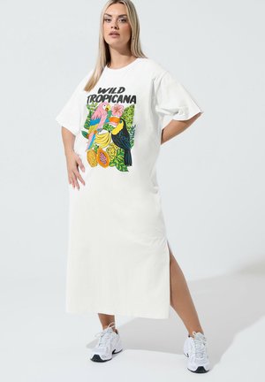 Kvinde iført hvid midi-kjole med tropisk fugle- og frugtprint og teksten "Wild Tropicana", kombineret med hvide sneakers, står selvsikkert.
