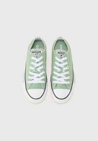 Grønne lerrets-sneakers med hvite snører, rund tå og hvit gummisåle med en svart aksentstripe og signaturmerkevare på tungen.