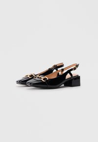 Zwarte lakleren slingback hakken met gouden kettingdetails, blokhakken en gespbanden, weergegeven op een effen witte achtergrond.