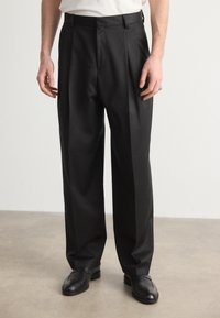 Schwarze, plissierte Hose aus glattem Stoff mit geradem Bein. Verfügt über Gürtelschlaufen und eine taillierte Passform. Trägt schwarze Schuhe.