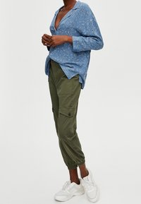 Femme portant un chemisier fleuri bleu à manches longues, un pantalon cargo vert olive avec poches, et des baskets blanches, se tenant les mains jointes.