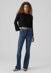 Pull court noir en tricot avec un motif texturé et des manches larges, associé à un jean en denim bleu évasé et des sandales noires à bout ouvert.