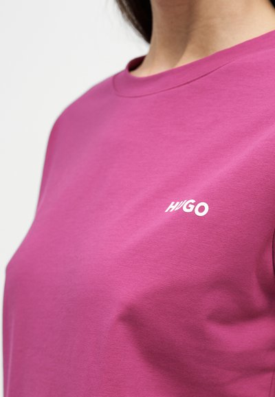 Gros plan d'une personne portant un t-shirt violet à col rond avec le nom de la marque Hugo imprimé en blanc sur la poitrine.