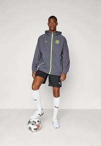 Gray hooded jacket with lime green accents, black shorts, knee-high white socks, and white athletic shoes. A football is at the model's feet.