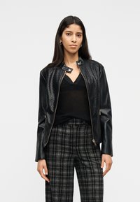 Γυναίκα με μακριά σκούρα μαλλιά που φοράει μαύρο υφαντό δερμάτινο μπουφάν, μαύρο μπλουζάκι με V λαιμόκοψη και ασπρόμαυρο καρό παντελόνι, στέκεται μπροστά σε λευκό φόντο.