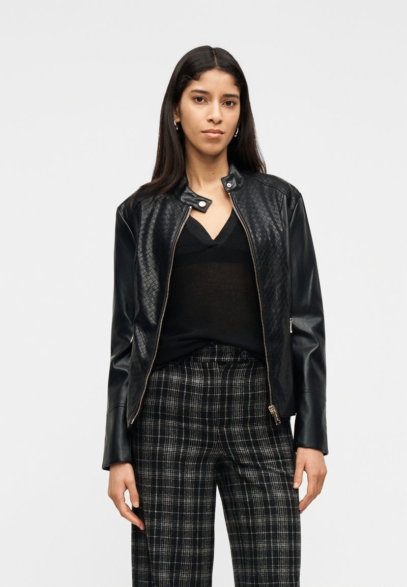 Γυναίκα με μακριά σκούρα μαλλιά που φοράει μαύρο υφαντό δερμάτινο μπουφάν, μαύρο μπλουζάκι με V λαιμόκοψη και ασπρόμαυρο καρό παντελόνι, στέκεται μπροστά σε λευκό φόντο.