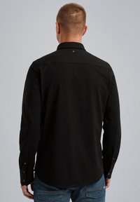 Czarna koszula z długim rękawem, z kołnierzykiem, z zapięciem na guziki przy mankietach oraz z małym detalem logo z tyłu, zestawiona z niebieskimi dżinsami.