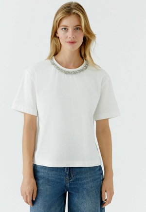 Wit katoenen T-shirt met korte mouwen, ronde halslijn, versierd met een zilveren decoratieve kraag. Draagt bij blauwe spijkerbroek.