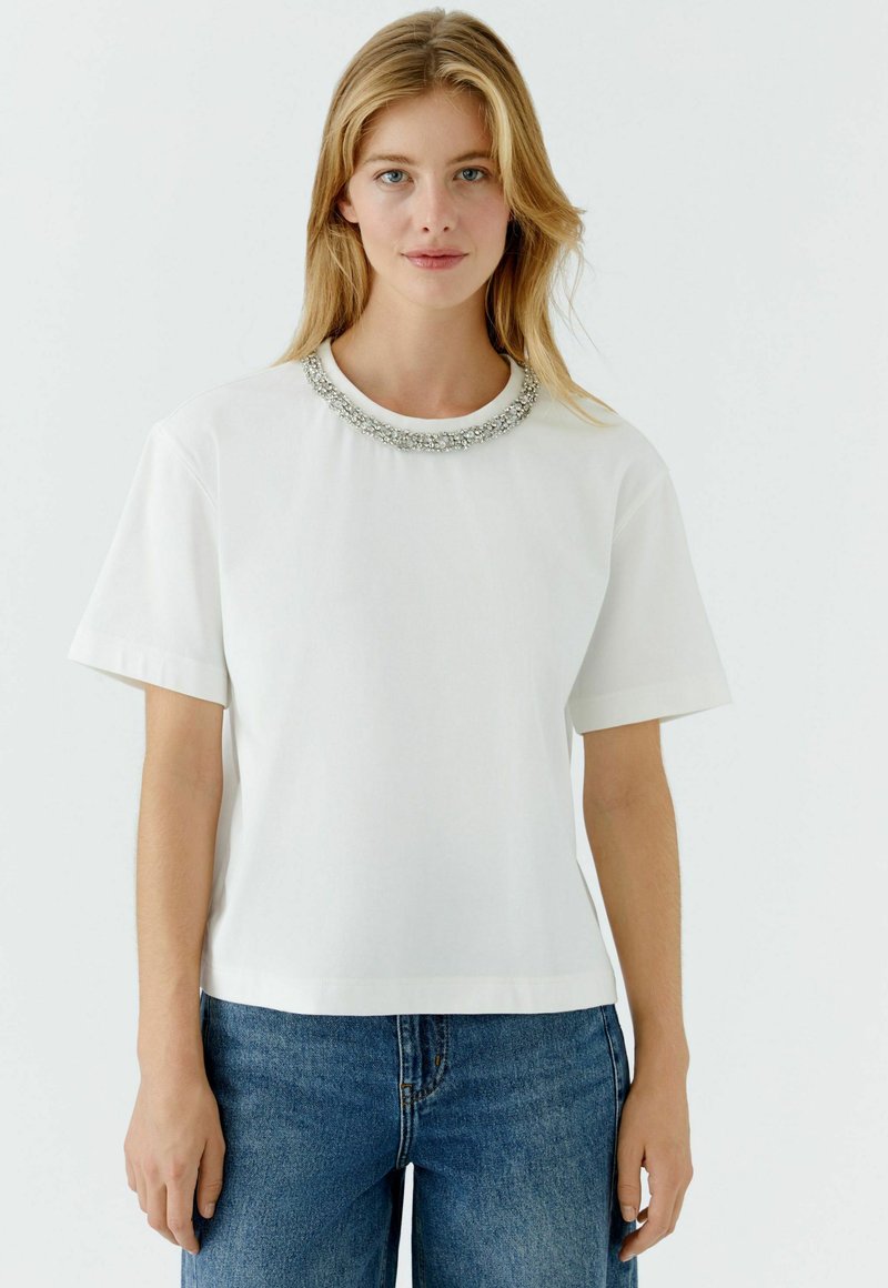 Weißes Baumwoll-T-Shirt mit kurzen Ärmeln und rundem Ausschnitt, verziert mit einem silbernen dekorativen Kragen. Kombiniert mit blauen Jeans aus Denim.