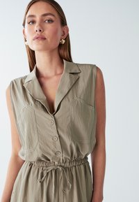 Robe chemise sans manches de couleur vert olive clair avec fermeture à boutons, col pointu, deux poches poitrine et taille à cordon. Tissu lisse et texturé.