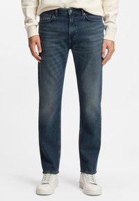 Vaqueros azul oscuro con acabado de mezclilla texturizada, corte recto, diseño de cinco bolsillos y sutil desgaste en los muslos. Combinados con zapatillas blancas.