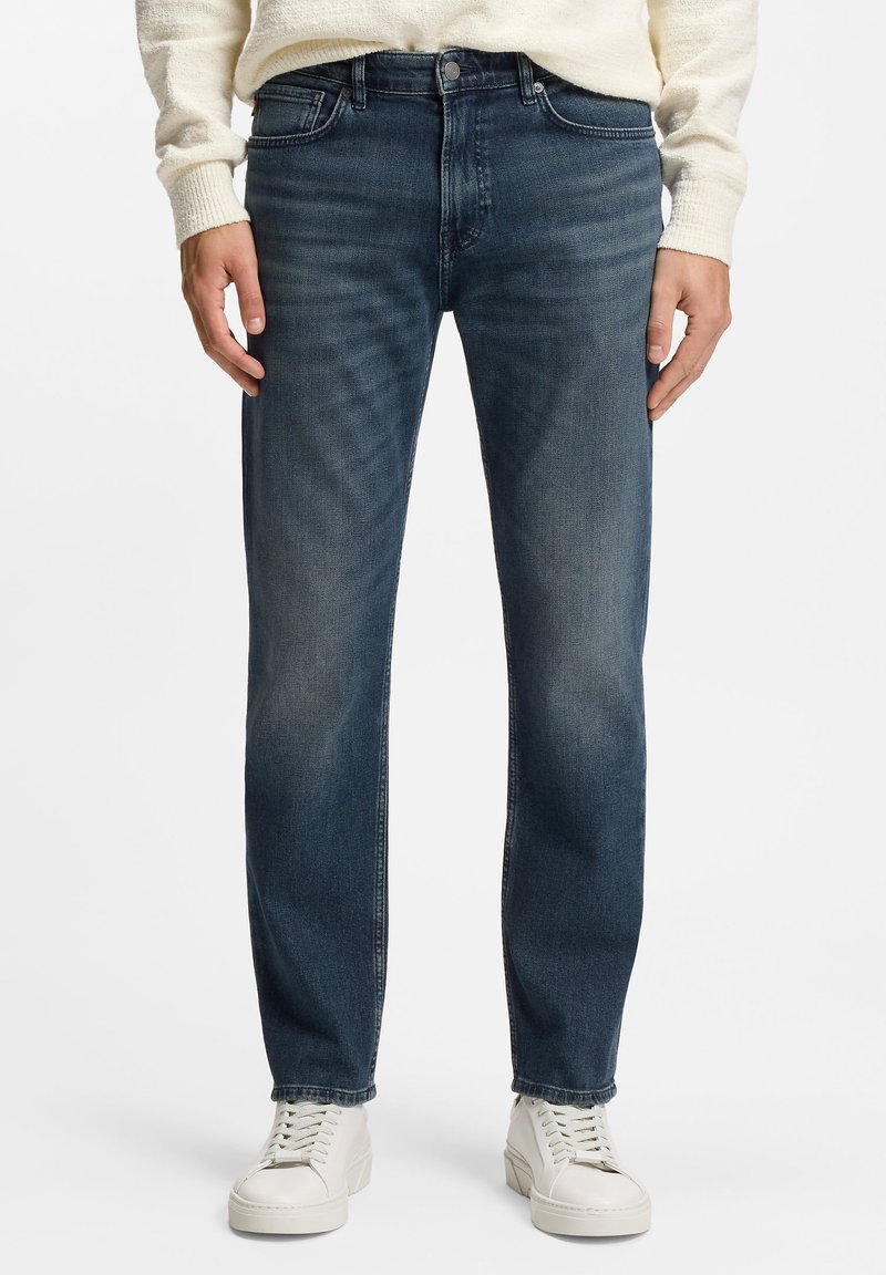 Vaqueros azul oscuro con acabado de mezclilla texturizada, corte recto, diseño de cinco bolsillos y sutil desgaste en los muslos. Combinados con zapatillas blancas.