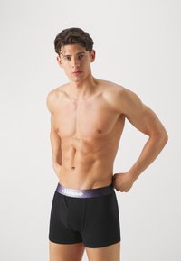 Schwarze Boxer-Shorts mit einem lila gebrandeten Bund, aus glattem Stoff hergestellt. Sie verfügen über ein enganliegendes Design und enden mittig am Oberschenkel.