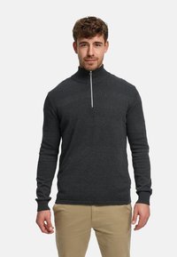 Mand med krøllet hår iført en mørkegrå tekstureret sweater med halv lynlås og beige bukser, stående foran en ensfarvet lys baggrund.
