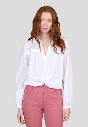 Blouse blanche à manches longues avec col boutonné, comportant des motifs de fleurs découpés sur les manches. Associée à un pantalon rose clair ajusté.