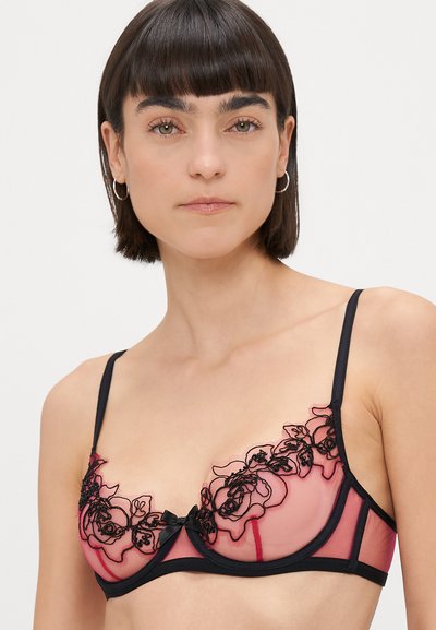 Sostén rosa transparente con bordado floral negro, soporte con aros y tirantes ajustables. Presenta un pequeño lazo negro en el centro.