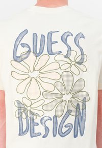 Cream-colored cotton t-shirt featuring a floral graphic in blue and olive green, with a mesh texture on some petals and bold text reading "GUESS DESIGN."