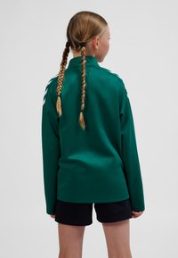 Grön långärmad tröja med krage och vita ränder på axlarna, kombinerad med svarta shorts, sedd bakifrån.