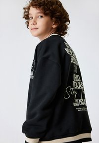 Svart sweatshirt med vit text och grafik. Har en avslappnad passform, ribbade muddarna och nederkant med kontrasterande ränder. Lockigt hår synligt.