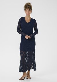 Navyblauwe kanten jurk met lange mouwen, een scalloped halslijn en doorzichtige onderkant. Het materiaal heeft een getextureerd bloemenpatroon.
