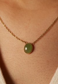 Gouden ketting met een ovale groene steen, gezet in een parelachtige gouden bezel, rustend tegen de huid. Ketting met gestructureerde schakels.