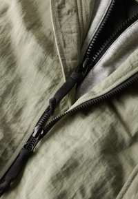 Olijf groene lichte jasstof met een subtiele textuur, voorzien van een zwarte rits en een verstelbaar koord. Binnenkant met zichtbare grijze stof.