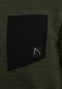 Tejido texturizado verde oscuro con un bolsillo de parche negro. El bolsillo tiene detalles del logotipo en blanco en la esquina inferior derecha.