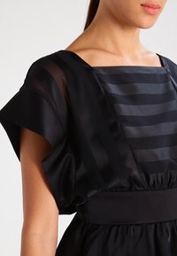 Sort kjole med firkantet halsudskæring, korte ærmer og et tekstureret design med skiftende ugennemsigtige og gennemsigtige striber. Elastisk taljedetalje.