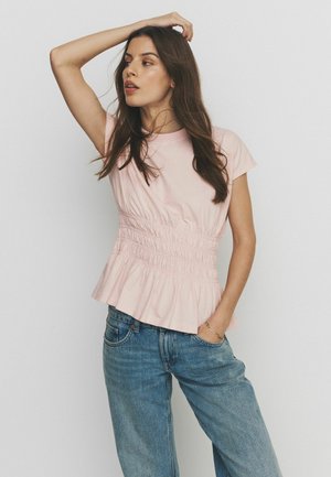 Pikkade pruunide juustega naine kannab heleroosat vöökohaga kogutud topi ja siniseid teksaseid, poseerides ühe käega peas ja teise taskus.