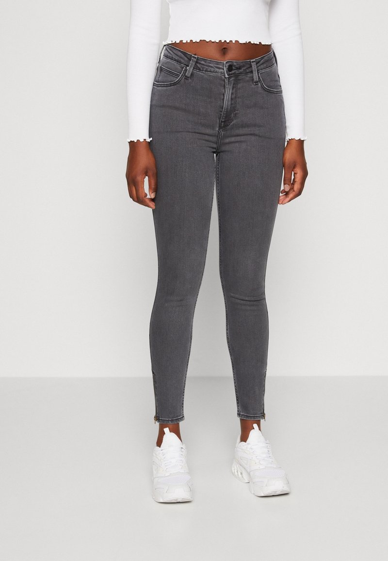 Femme portant un jean skinny gris foncé avec des fermetures éclair à la cheville, un haut blanc à manches longues court, et des baskets blanches, debout devant un fond clair uni.